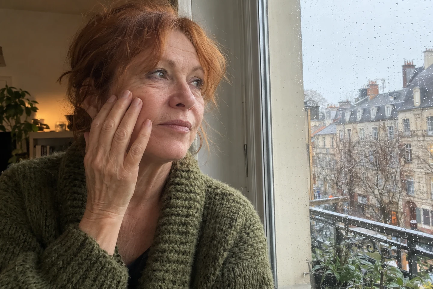 Une femme observe sa joue près d’une fenêtre un jour de pluie