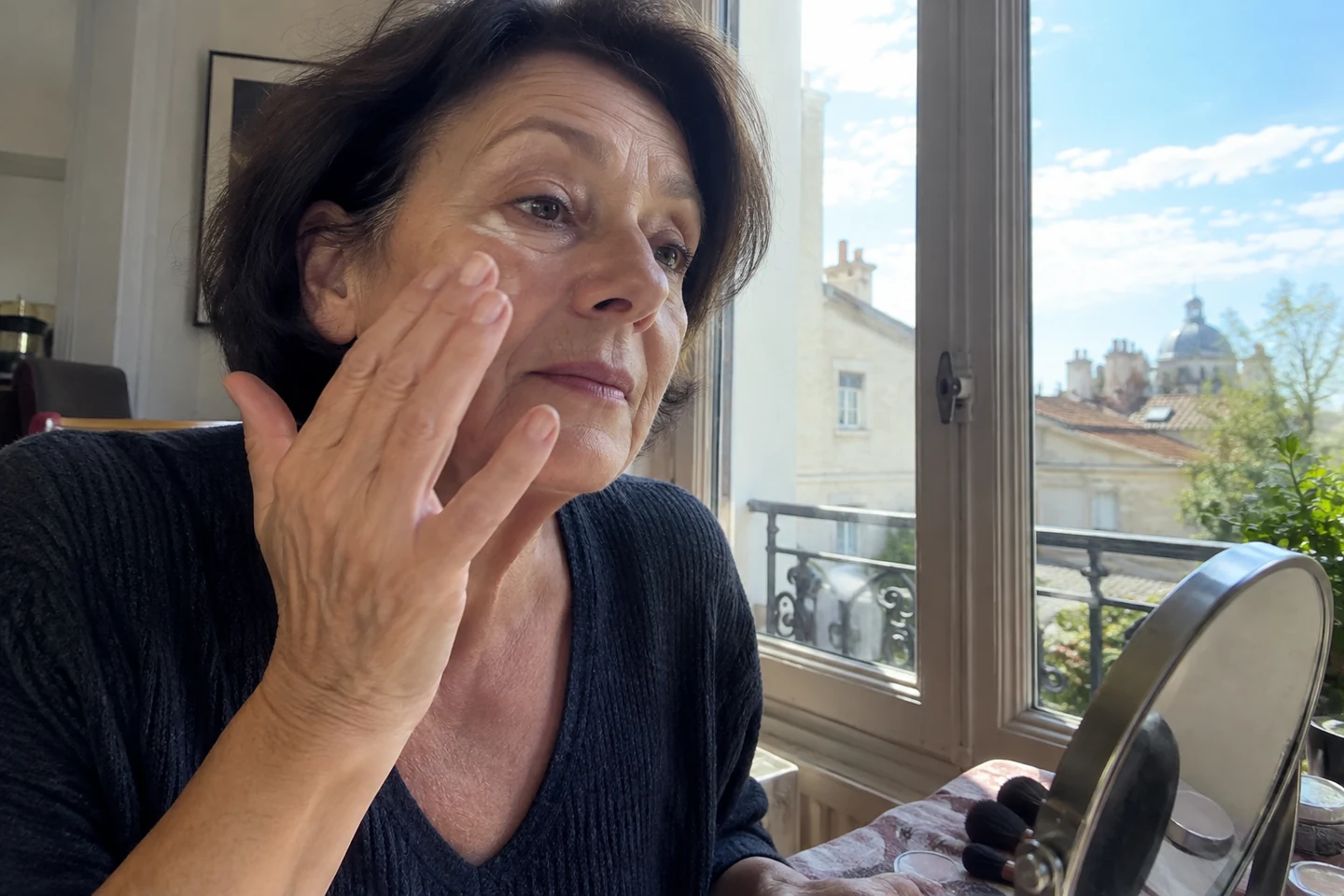 Une femme ajuste une couche légère de maquillage près d’une fenêtre
