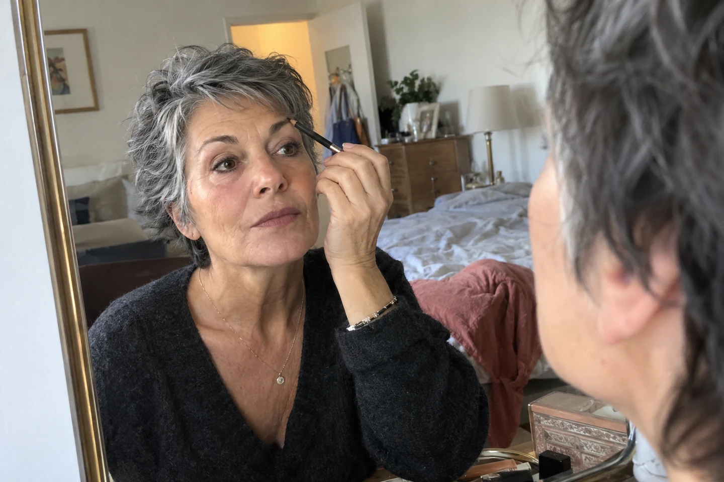 Une femme aux cheveux courts gris ajuste ses sourcils devant un miroir