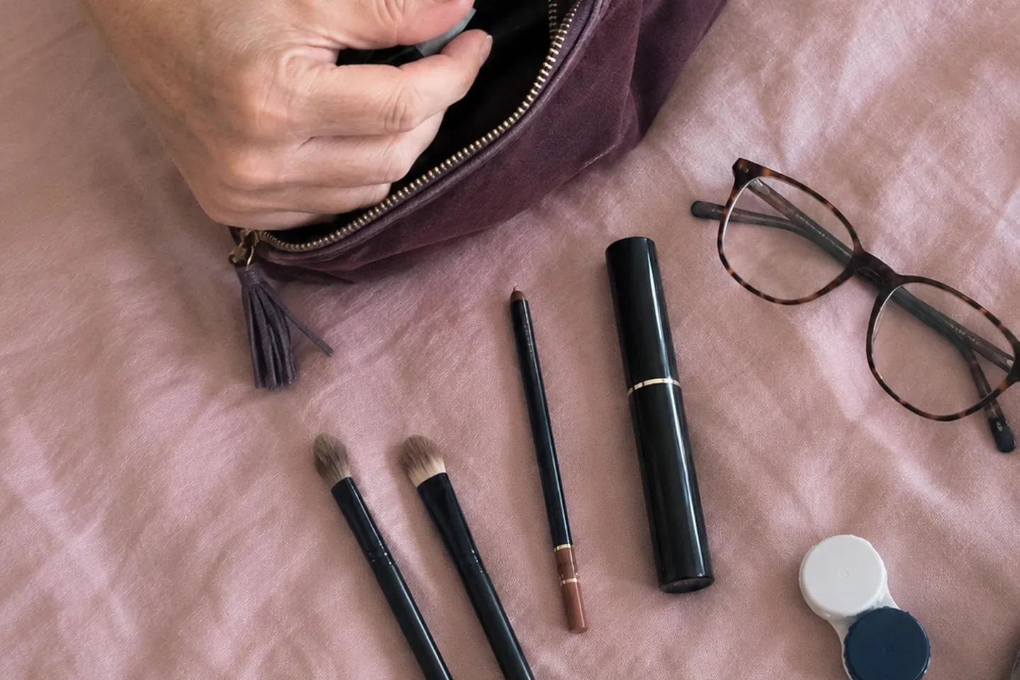 Mains rangeant une trousse avec du maquillage, des pinceaux et des lunettes sur un linge rose