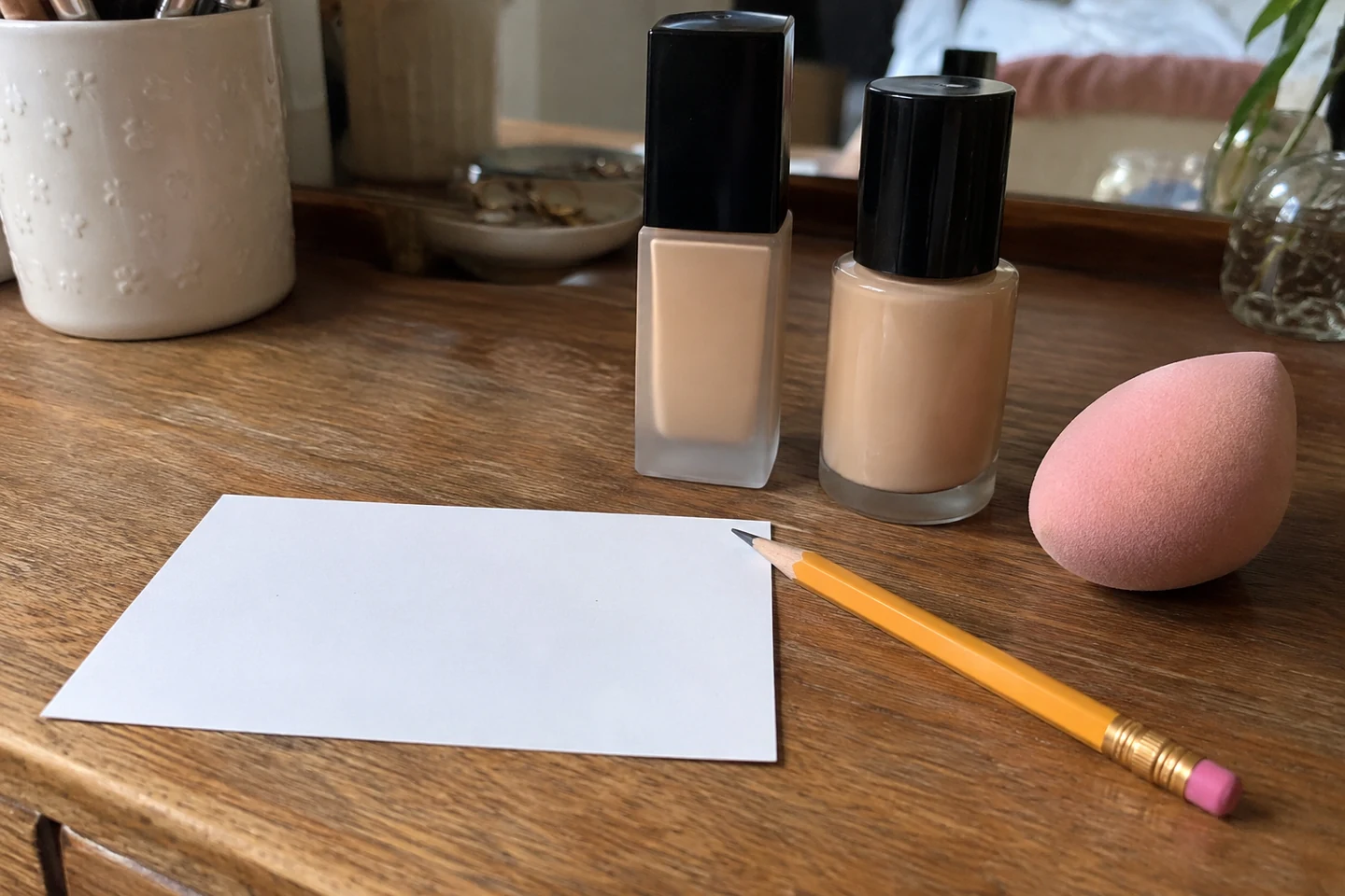 Deux fonds de teint fermés, une éponge rose, un crayon et une carte vierge sur une table.